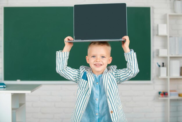 Niedlicher kleiner Schuljunge mit Laptop auf dem Kopf in einem Klassenzimmer
