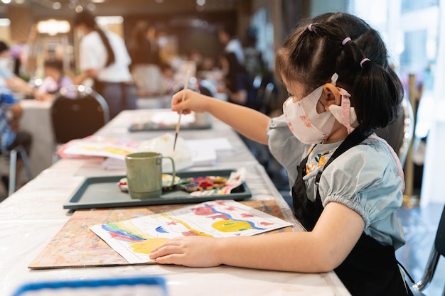 Niedliches kleines asiatisches Baby mit Maske, das lächelnd mit bunten Farben mit Aquarell malt Asiatisches Mädchen mit Pinselzeichnungsfarbe Baby-Aktivitäts-Lifestyle-Konzept Konzept der sozialen Distanz