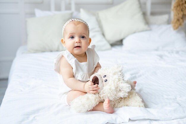 Niedliches kleines Baby in weißem Baumwollbody, das lächelt oder lacht und mit einem Teddybär-Mädchen auf einem hellen Bett zu Hause im Kindergarten in der Nähe spielt