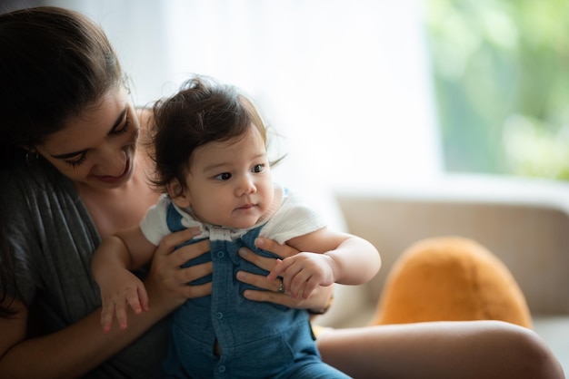 Niedliches kleines Kleinkind, das spielt und genießt, während es sich in den Armen der jungen Mutter zu Hause umarmt