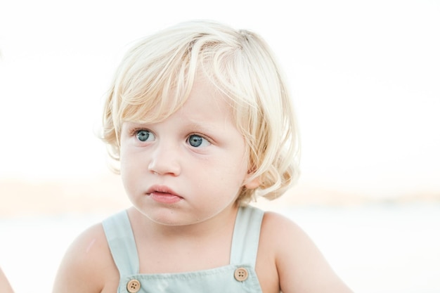 Niedliches kleines Kleinkind mit blonden Haaren, das während der Sommerferien vor verschwommenem Hintergrund auf das Meer blickt