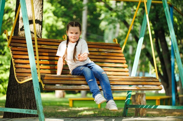 Niedliches kleines Mädchen, das auf einer Holzschaukel auf dem Spielplatz im grünen Park sitzt Sommerferien im Touristenzentrum des Lagers Gehen und Spielen im Freien Sportaktivitäten und gesunde Lebensweise Erholung