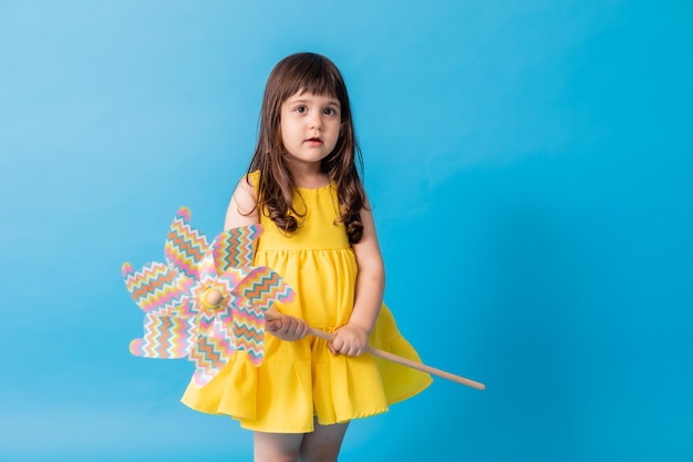 Niedliches kleines Mädchen in einem gelben Kleid auf blauem Hintergrund hält eine bunte Kinderspielzeug-Windmühle in der Hand