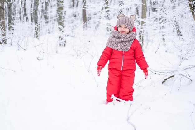 Niedliches kleines Mädchen in rosa Schneeanzug und Strickmütze und Schal spielt mit Schnee im Winterwald