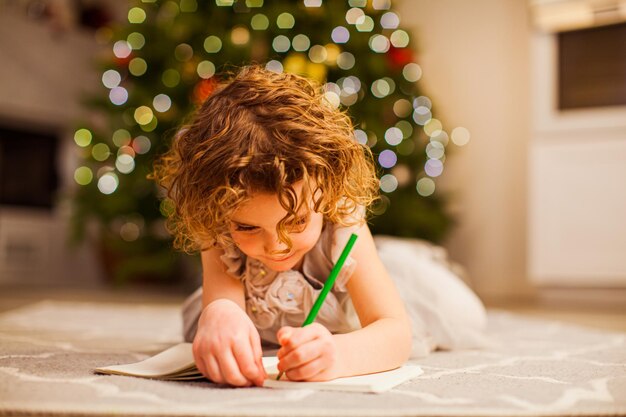 Niedliches kleines Mädchen mit lockigem Haar in schönem Kleid, das auf einem Boden liegt und in ihrem Notizbuch mit grünem Bleistift schreibt Weihnachtsbaum mit Lichtern auf verschwommenem Hintergrund