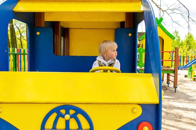 Niedliches, lustiges, blondes, kleines Kleinkind, Junge, der in einem Spielzeugautobus auf dem Spielplatz sitzt und fährt. Körperlich-emotionale Entwicklung der Kinder und Kindergartenkonzept für Kindertagesstätten