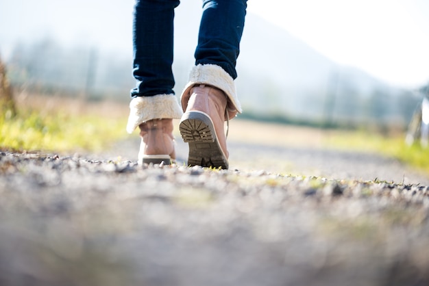 Niedrige Bodenansicht mit flachem Dof der Füße einer Frau in Jeans und knöchelhohen Lederstiefeln, die auf einem ländlichen Weg abseits der Kamera spazieren gehen.