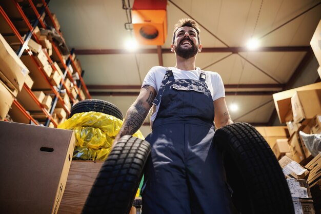 Niedrige Winkelansicht des lächelnden fleißigen Arbeiters in Overalls, die Reifen tragen und im Lager in Import- und Exportfirma gehen.