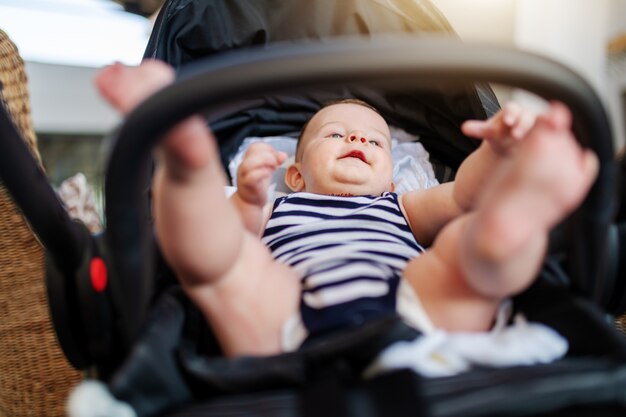 Niedrige Winkelansicht des verspielten kaukasischen glücklichen Jungen 6 Monate alt gekleidet in gestreifter Babykleidung, die im Kinderwagen sitzt und nach oben schaut.