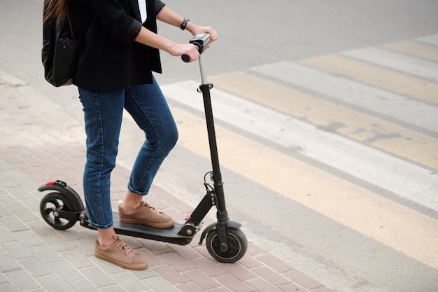 Niedriger Abschnitt der jungen Frau in der blauen Jeans und in der schwarzen Jacke, die ein Bein auf Elektroroller halten, während sie mit Zebrastreifen auf der Straße stehen