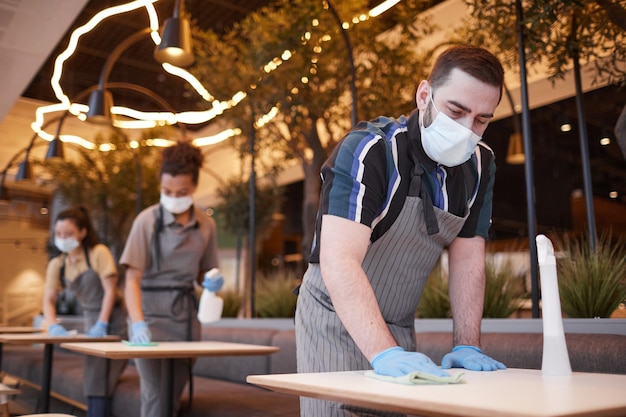 Niedriger Blickwinkel auf Arbeiter, die Masken in Reihe tragen, während sie Tische im Café-Innenraum reinigen, kovides Sicherheitskonzept, Kopierraum