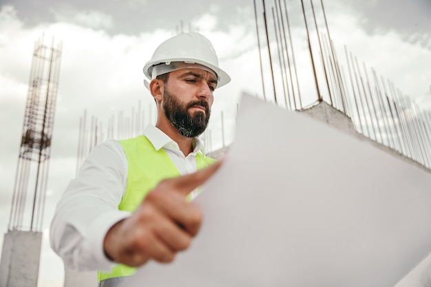 Niedriger Winkel des konzentrierten erwachsenen bärtigen männlichen Ingenieurs im Helm und in der Weste, die auf Baustelle stehen und Blaupause untersuchen