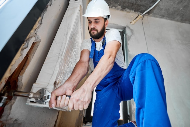 Foto niedriger winkel eines bärtigen jungen mannes in sicherheitshelm mit verstellbarem schraubenschlüssel bei der installation eines heizkörpers fokus auf den händen konzept der heizkörperinstallation klempnerarbeiten und heimrenovierung