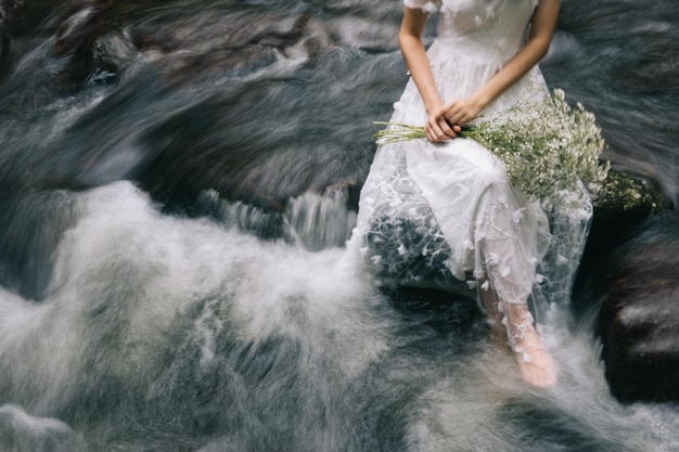 Niedriges Teil einer Frau in Hochzeitskleidung sitzt auf einem Felsen über dem Fluss