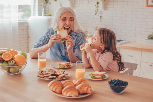 Nieta y abuela comiendo sándwiches para desayunar sentados en la mesa de la cocina