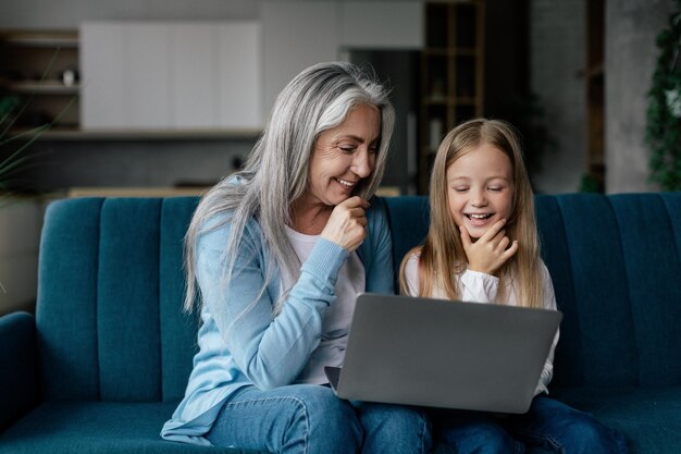La nieta caucásica alegre y la abuela mayor piensan ver un video en un estudio de computadora en línea