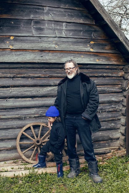 Nieto con sombrero azul y botas con un abuelo en un perchero de piel de oveja en un cobertizo de madera en la aldea en primavera en Rusia