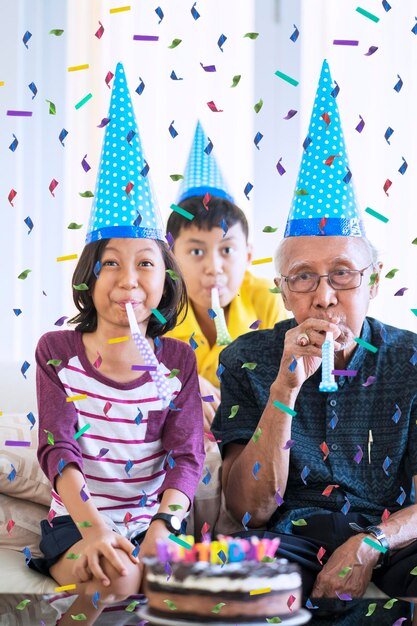 Nietos felices celebrando el cumpleaños de su abuelo mientras usan sombrero de cumpleaños con confeti cayendo en casa