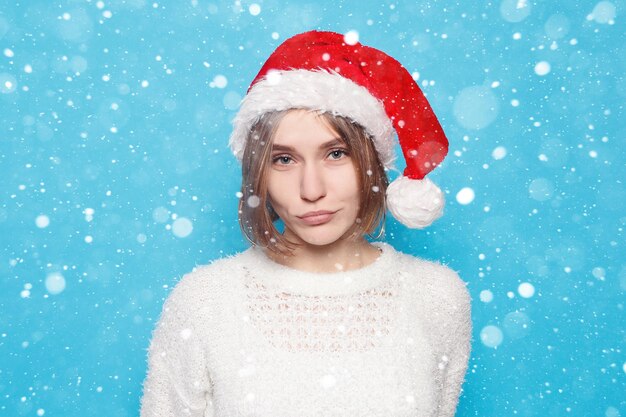 Nieve, invierno, Navidad, gente, concepto de belleza - bastante rubia con sombrero de Navidad sobre fondo azul claro de nieve. Retrato de joven hermosa linda chica alegre sonriendo mirando a la cámara