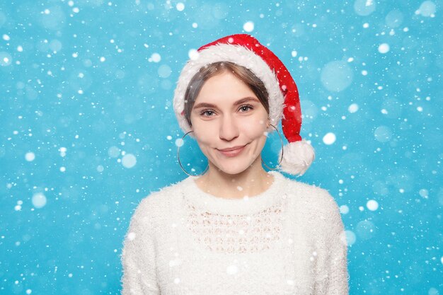 Nieve, invierno, Navidad, gente, concepto de belleza - bastante rubia con sombrero de Navidad sobre fondo azul claro de nieve. Retrato de joven hermosa linda chica alegre sonriendo mirando a la cámara