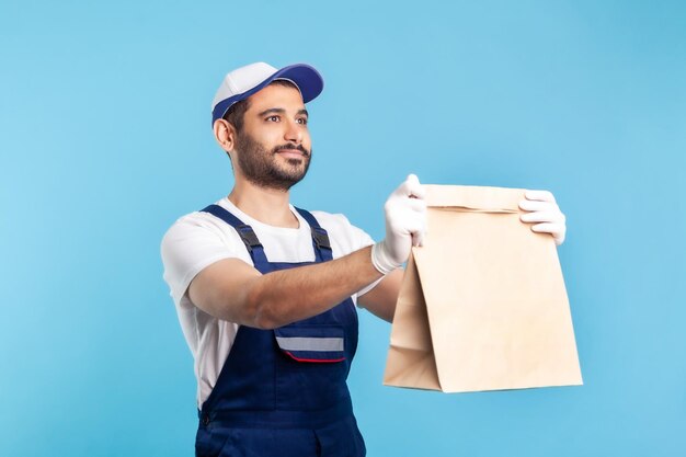 Nimm es. Porträt eines fröhlichen bärtigen Handwerkers in Overalls und Handschuhen, der bestellte Pakete gibt. Kurier liefert Lebensmittel in Papiertüten, Postdienste. Studioaufnahme im Innenbereich isoliert auf blauem Hintergrund