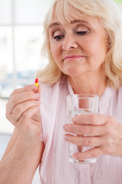 Nimmt ihr täglich Medikamente. Depressive ältere Frau, die Pille und ein Glas Wasser hält, während sie drinnen sitzt