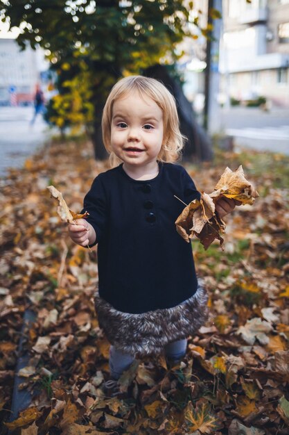 Una niña de 1,5 años está de pie y sonriendo. El niño lanza hojas. Chica alegre en el bosque de otoño