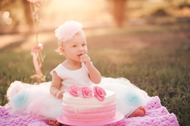 Niña de 1 año comiendo pastel de cumpleaños cremoso sentada en la hierba verde con globos rosados al aire libre