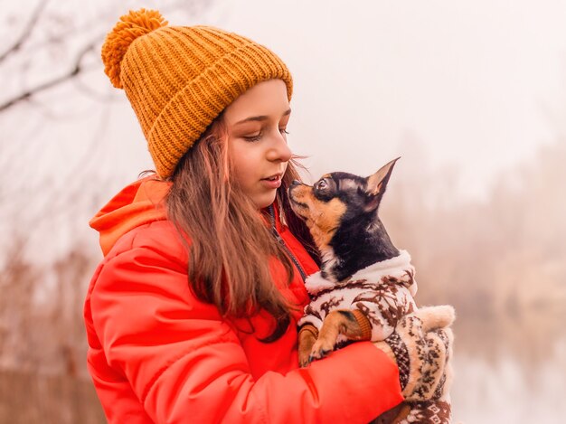 Una niña de 10-11 años con una chaqueta naranja con su mascota, un perro Chihuahua, en la naturaleza.