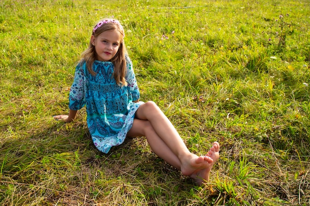 Una niña de 10 años con un vestido azul sentada en un campo de hierba y  flores, piernas largas y desnudas, descalza