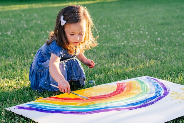 Niña de 2-4 años pinta el arco iris y el sol en una hoja grande de papel, sentada en el césped verde