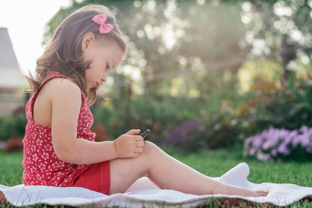 Una niña de 3 a 4 años con ropa roja se sienta en una manta sobre hierba verde y mira el teléfono móvil. Niños, usando aparatos