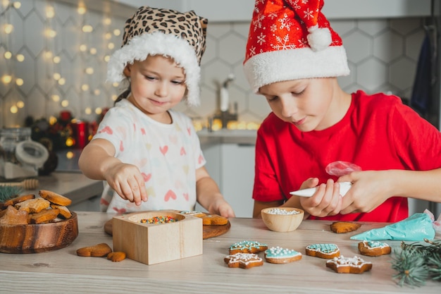 Niña de 3 años y niño de 8 años con gorro de Papá Noel decoran galletas de jengibre con glaseado. Hermanos en la cocina de Navidad