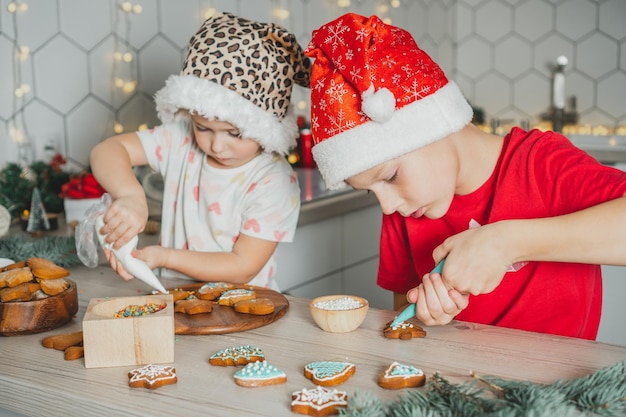 Niña de 3 años y niño de 8 años con gorro de Papá Noel decoran galletas de jengibre con glaseado Hermanos en la cocina de Navidad