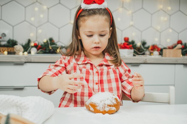 Niña de 3 años en rojo gorro de Navidad y camisa a cuadros prepara la masa para galletas de jengibre en la cocina