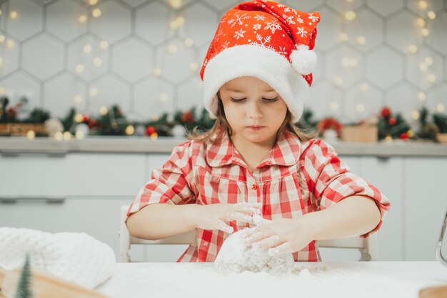 Niña de 3 años en rojo gorro de Navidad y camisa a cuadros prepara la masa para galletas de jengibre en la cocina