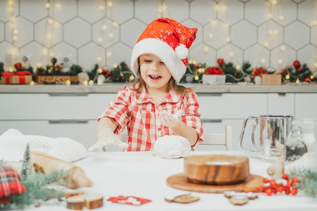 Niña de 3 años en rojo gorro de Navidad y camisa a cuadros prepara la masa para galletas de jengibre en la cocina