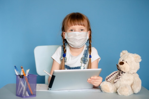 Una niña de 5 a 6 años sentada en una mesa realiza tareas remotas en la tableta. Aislar en una pared azul. Coronavirus, niño enmascarado.