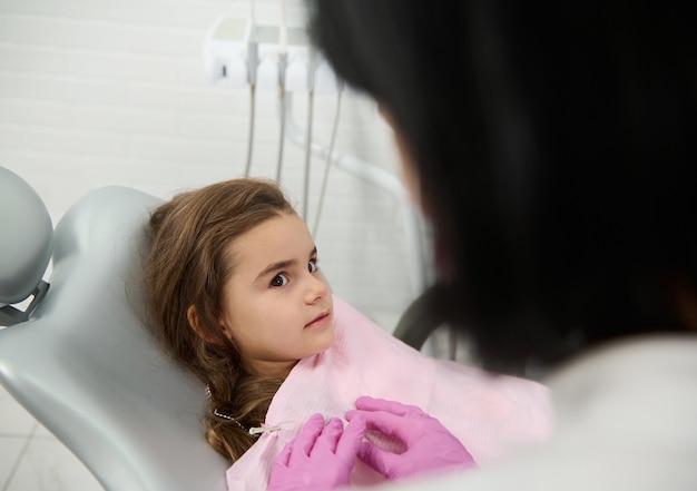 Niña de 5 años en silla de dentista mirando higienista preparándola para el chequeo durante la cita médica en la clínica de odontología. Concepto de diagnóstico temprano de caries de dientes de leche. Práctica dental para niños