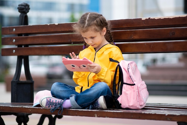 Una niña de 6-7 años con tablet PC en sus manos. Ella se sienta afuera en un banco. Publicidad de nuevo gadget, aplicación educativa. Juegos. Los niños usan la tecnología.