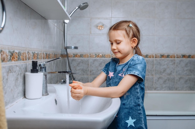 Una niña de unos 6 años con un vestido estampado de mezclilla se para en un fregadero blanco con agua corriente y se lava las manos de los gérmenes