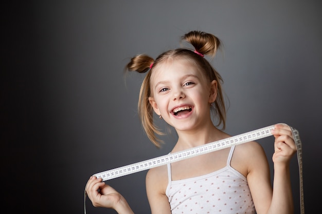 Niña de 7-8 años riendo, niño con una cinta métrica (centímetro) en una pared gris