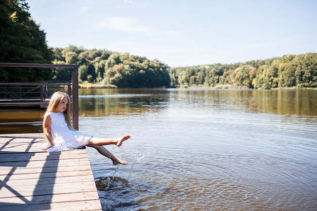 Foto una niña de 7 años con cabello largo y rubio junto al lago se sienta en un embrague con las piernas en el agua. ella salpica sus pies en el lago. chica descalza con un vestido blanco con pelo largo.