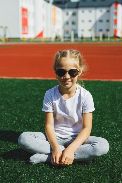 Una niña de 78 años con una camiseta blanca y gafas de sol está sentada en el campo de deportes La niña está sentada en la hierba verde en un día soleado