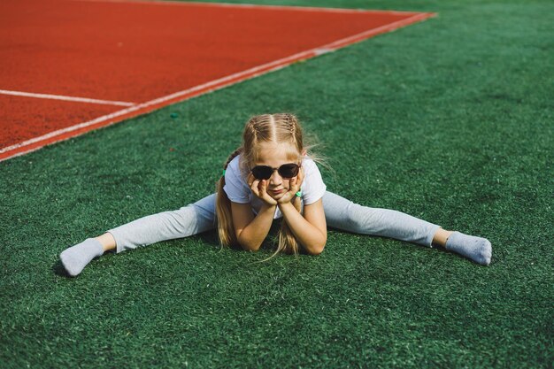 Una niña de 78 años con una camiseta blanca y gafas de sol está sentada en el campo de deportes La niña está sentada en la hierba verde en un día soleado