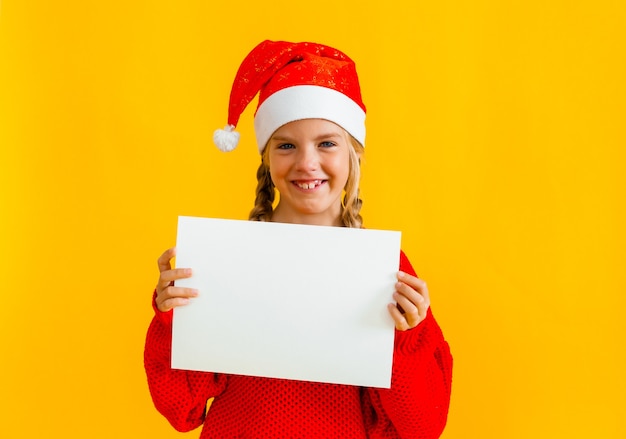 Una niña de 8 años con cabello rubio con un gorro de Papá Noel sostiene una hoja de papel blanca sobre un fondo amarillo.
