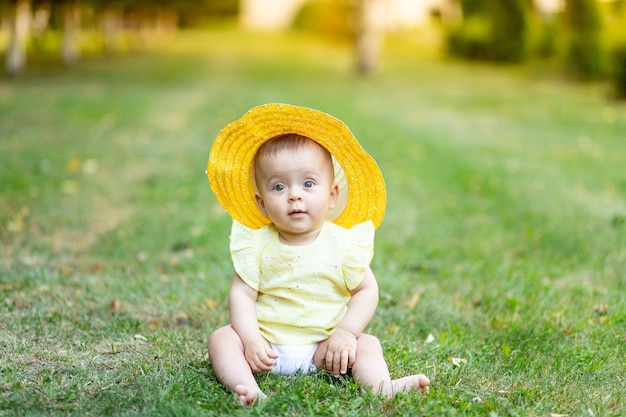 Una niña de 8 meses está sentada en el verano sobre la hierba verde con un vestido amarillo de verano y un sombrero