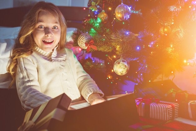 La niña abre la caja de luz cerca del árbol de Navidad. tarde noche