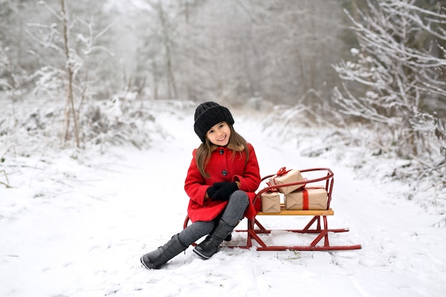 Una niña con un abrigo está sentada en un trineo con regalos de Navidad en un invierno nevado