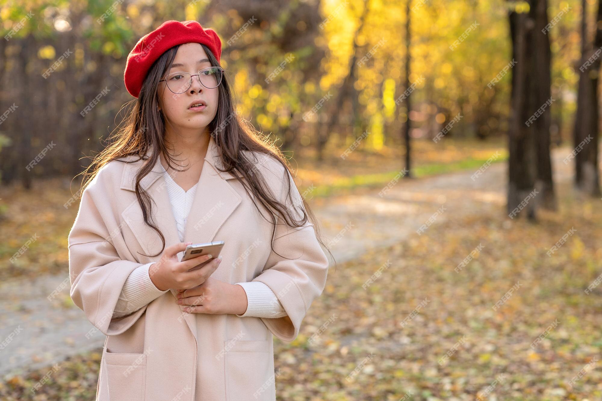 Una niña con un abrigo ligero y una boina roja camina en el parque de otoño sostiene un teléfono en la mano. | Foto Premium